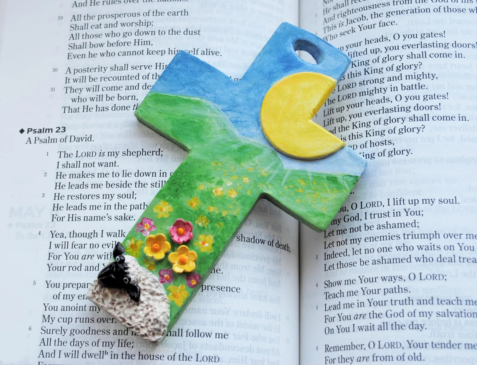 An air-dry clay cross with a blue sky, a yellow moon, green fields, spring flowers and a sheep, lying on an open Bible at Psalm 23