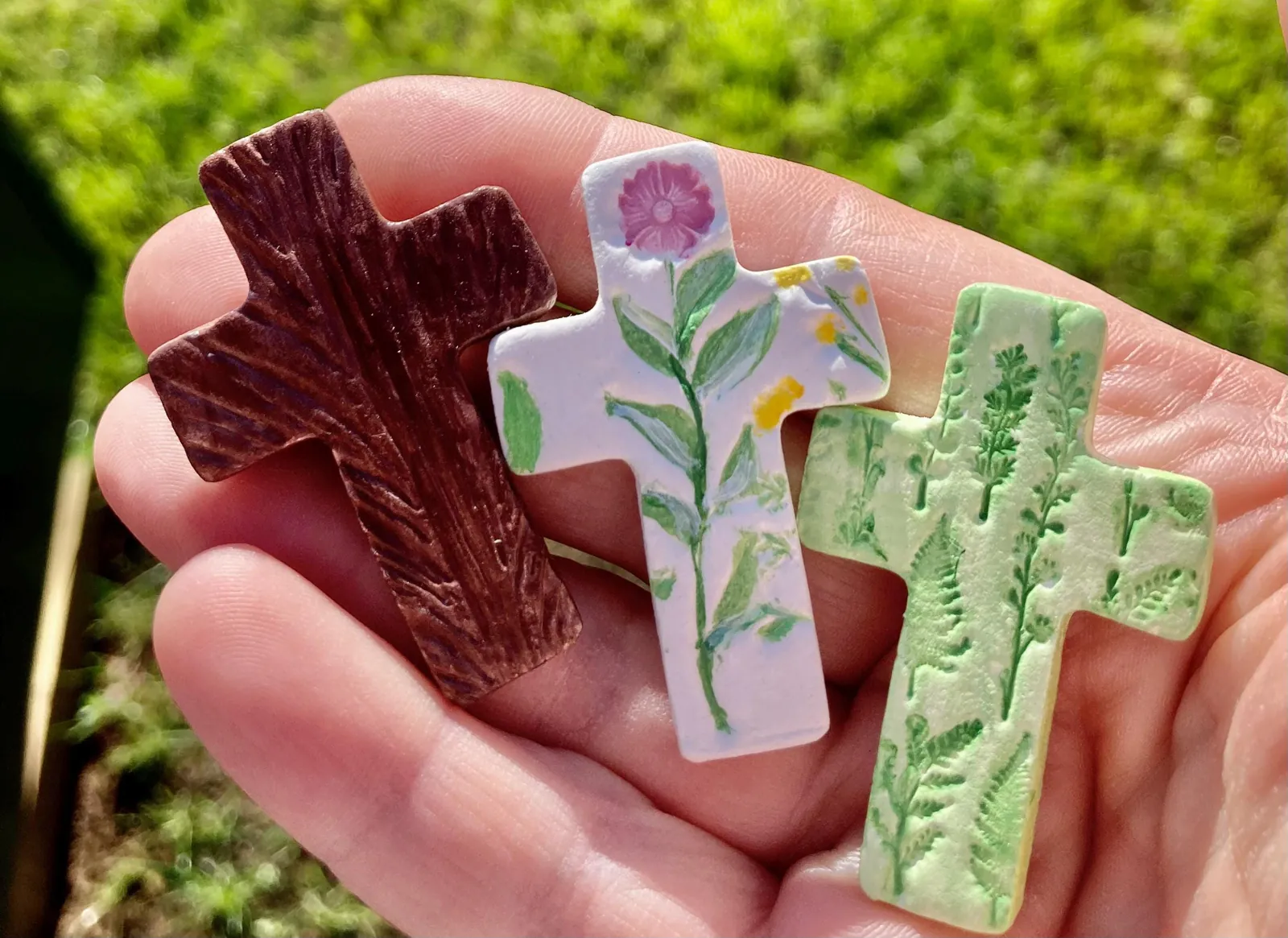 Three small air-dry clay crosses held in the palm of a hand: one brown with a wood-grain texture, one white with pink and yellow flowers, one green with fern and leaf prints